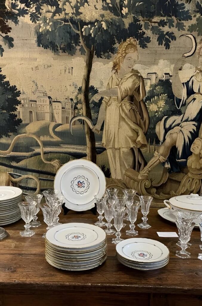 Delicate French china dishes and crystal glasses displayed in front of an antique woven tapestry.