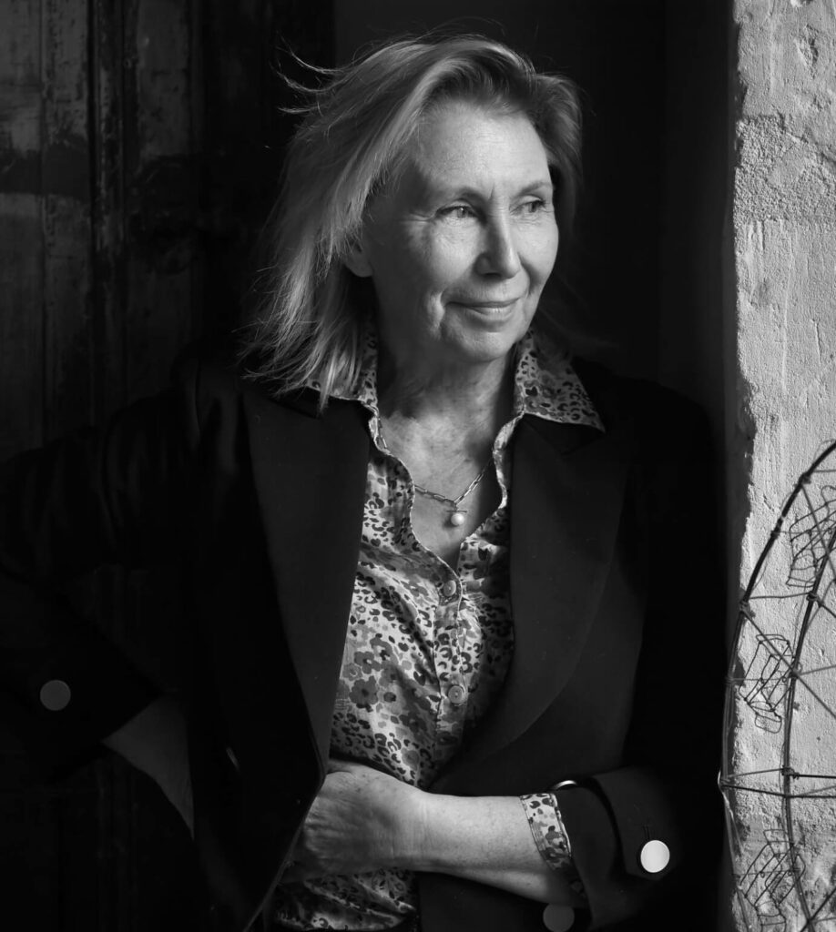 Sharon Santoni smiling and leaning on a rustic sone wall.