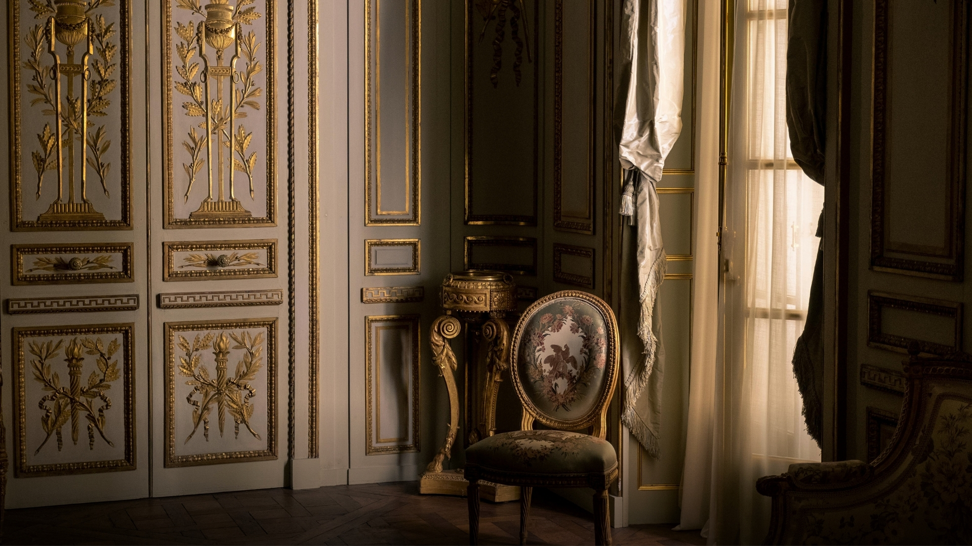 An ornate Louis XVI-style interior with gold-leaf doors, a round backed embroidered chair, and sweeping curtains.