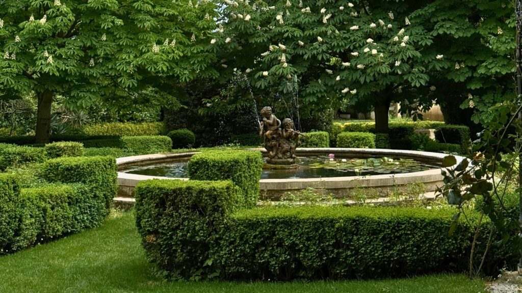 A lush green provincial garden with a stone pond, floating water lilies, and decorative cherub statues.