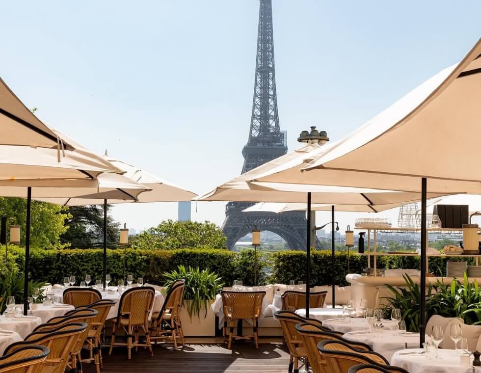 Wicker chair lined tables shaded by umbrellas at the Girafe rooftop restaurant, facing the Eiffel Tower.