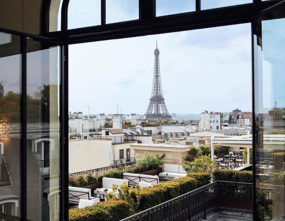 View of the Eiffel Tower through a window on a bright summer day, with a rooftop below adorned with greenery and seating.