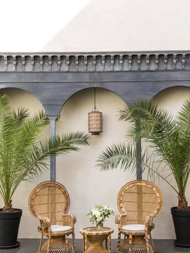On a Moroccan terrace, wicker chairs are framed by wooden arches and trees.