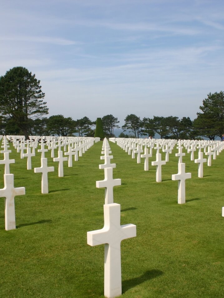 Vast rows of white headstones commemorating fallen soldiers at the Normandy American Cemetery, facing a tree-lined border.