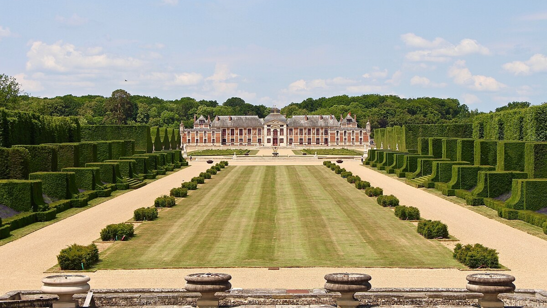 A sprawling Normandy estate accompanied by a garden lined with square hedges and trimmed shrubs.