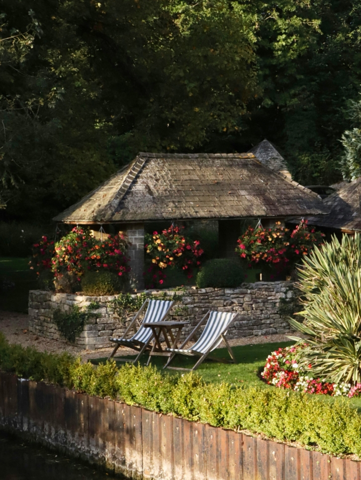 Lush garden in the Cotswolds with blue and white chairs overlooking water.