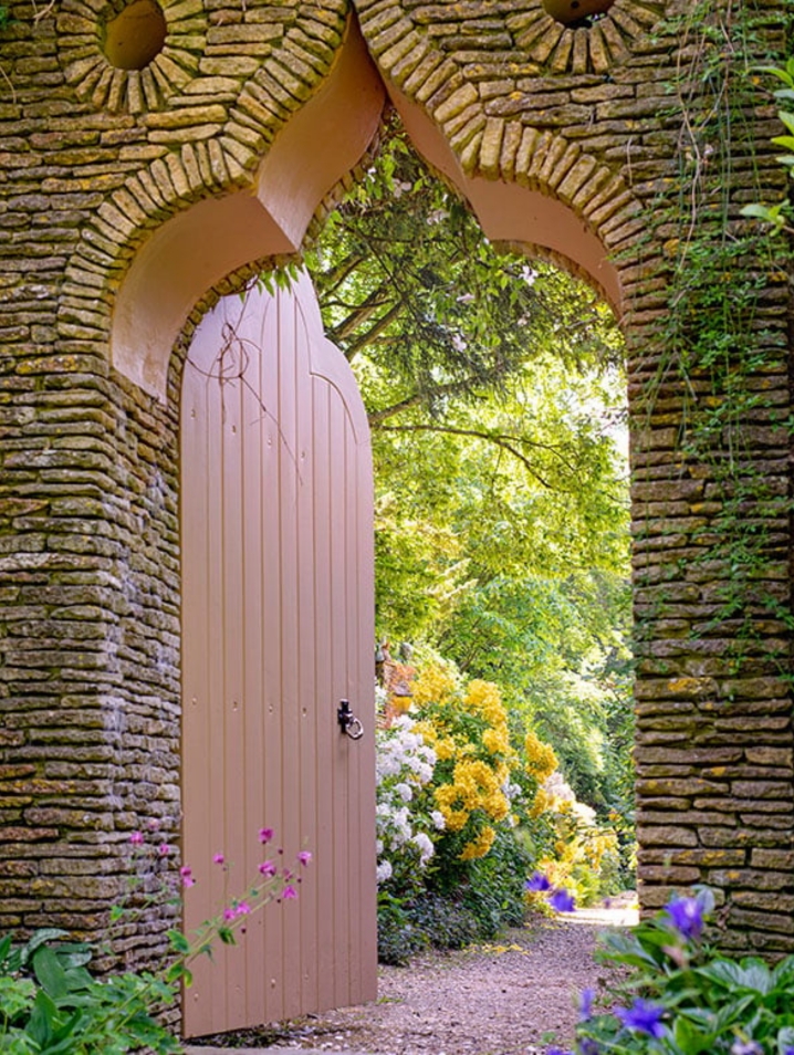 Brown door set in a stone wall leading to a flower garden.