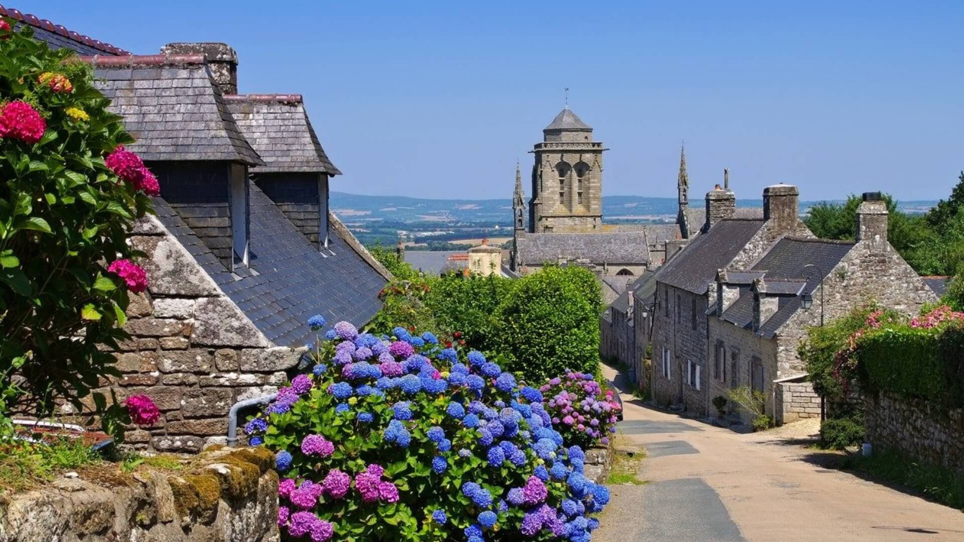 Blue hills frame the village of Locronan with its steeple and a flower-lined central walking path.