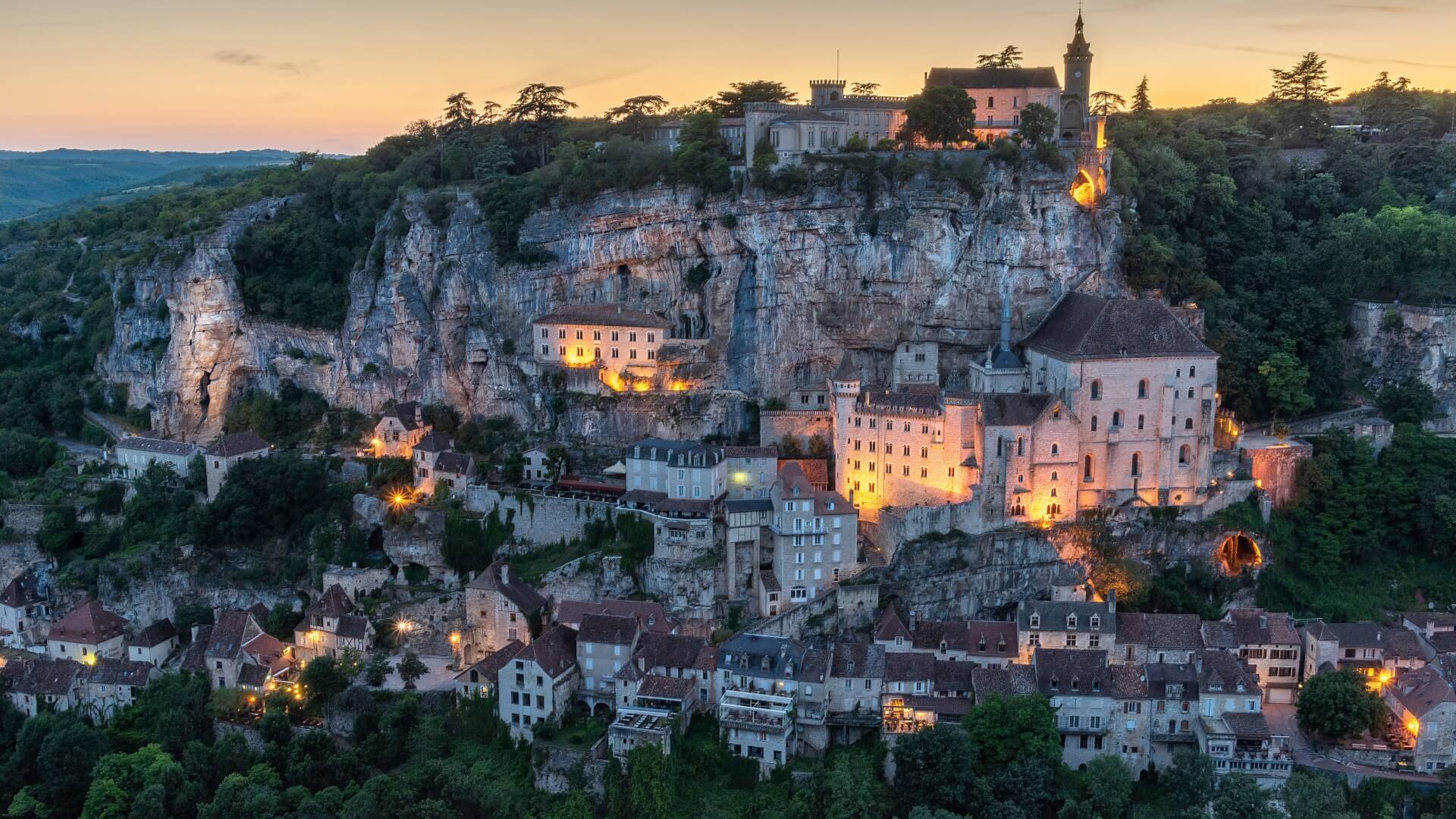The glowing, rugged, cliffside village of Rocamadour, clustered with historic stone homes, at twilight.