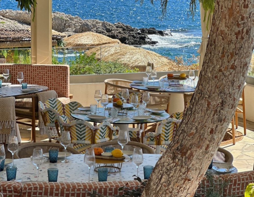 La Pinède’s shaded dining area with blue and yellow accents overlooks the rocky Mediterranean coast.