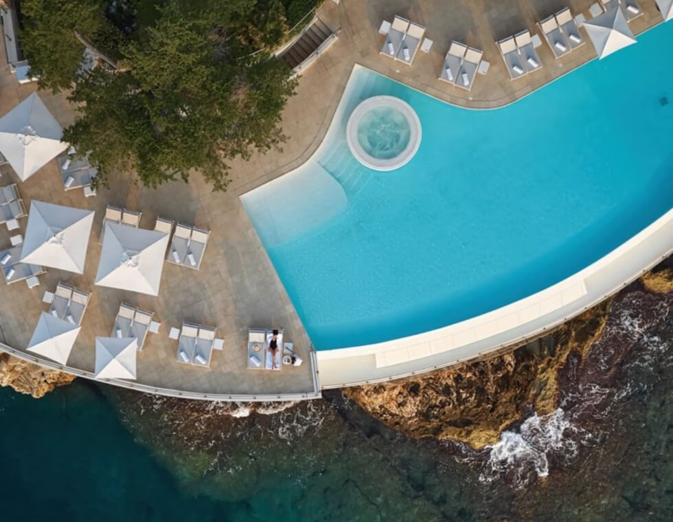 Bird's eye view of Cap Estel's aquamarine luxury swimming pool, framed by lounge chairs and umbrellas overlooking the Mediterranean Sea.
