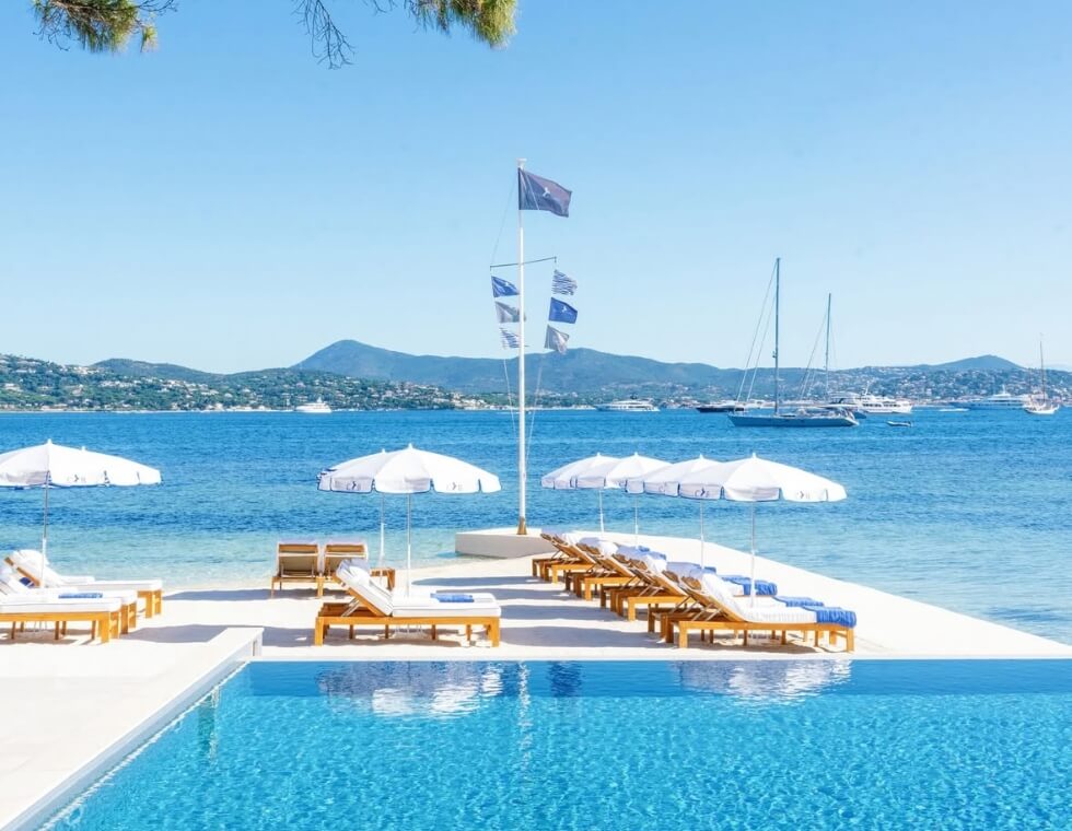 Cheval Blanc's rich, blue, swimming pool is perched seaside, framed by lounge chairs, mountains, and sailboats.