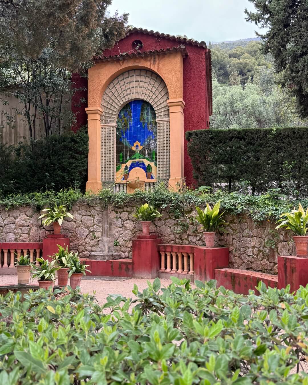 French Riviera garden with blooming potted plants, ocher benches, stone walls, and a framed mosaic.