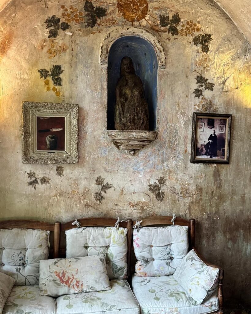 An ancient frescoed wall decorated with framed photos and a religious statue, accompanied by an antique white chaise.