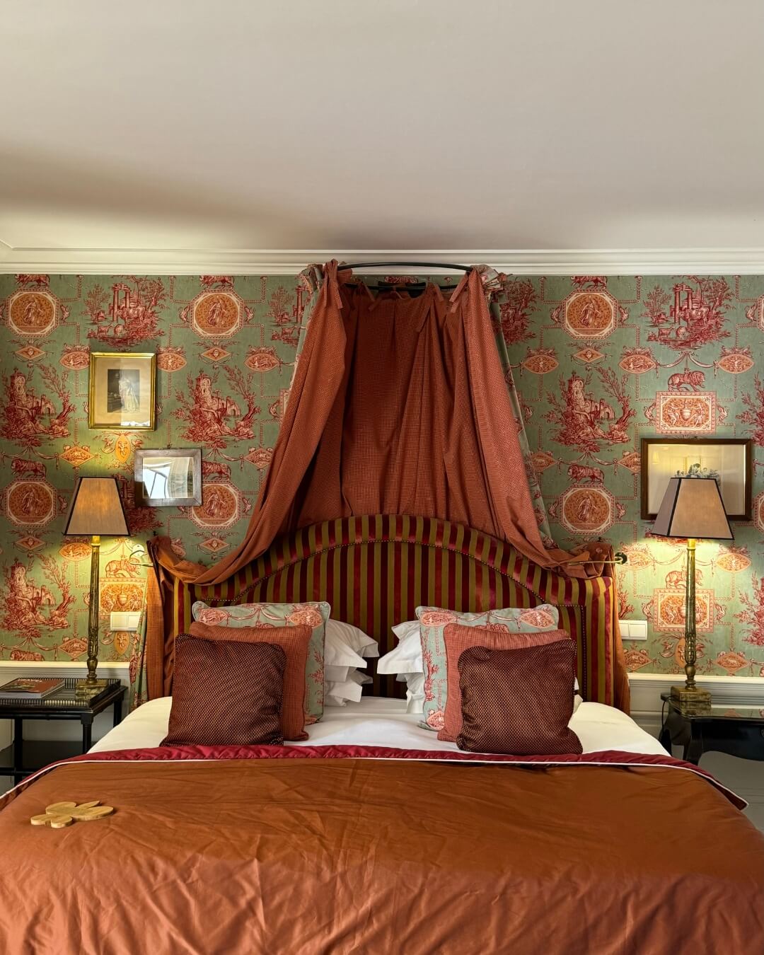 French Riviera boutique hotel bedroom with a made bed, warm accents, canopy frame, and teal patterned wallpaper.