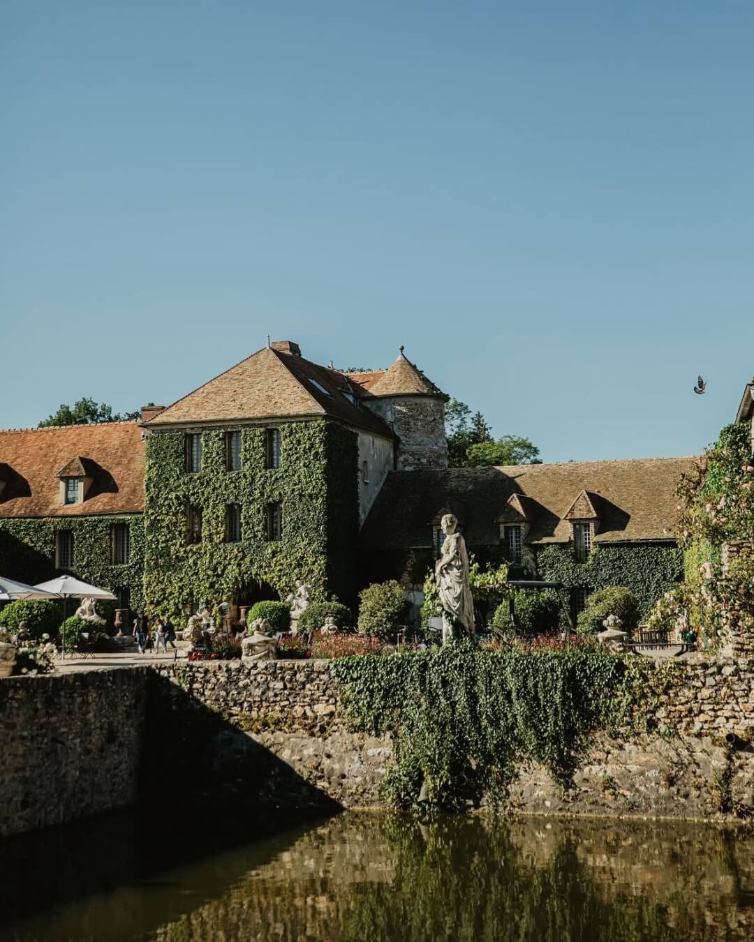 The Maison de Campagne estate is covered in lush ivy, featuring a garden and pond framed by stone walls.