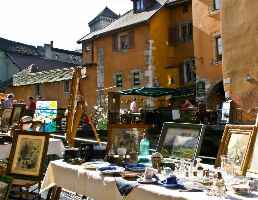 Table at Annecy outdoor antique market with framed paintings, glassware, and unique plates.
