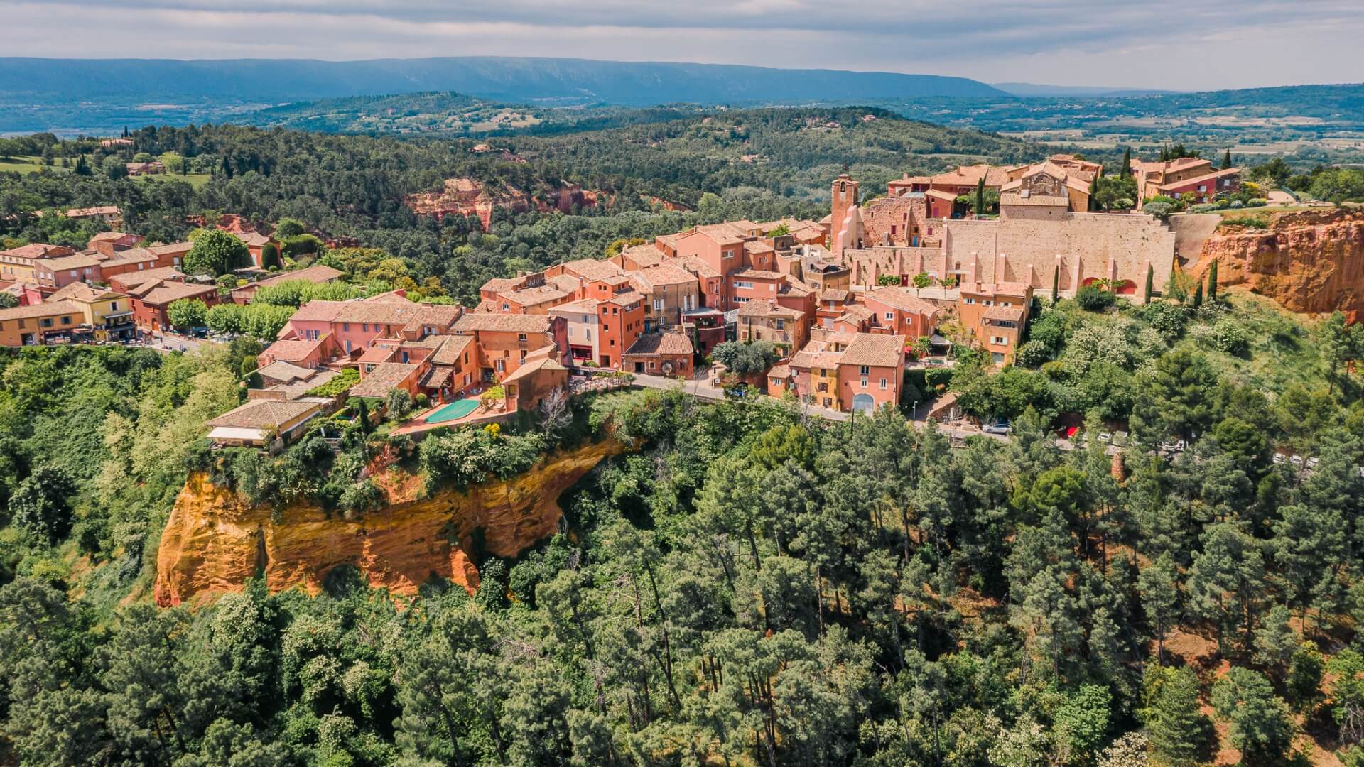 Red hued cliffside village of Roussillon in Provence, France, surrounded by hills and forest.