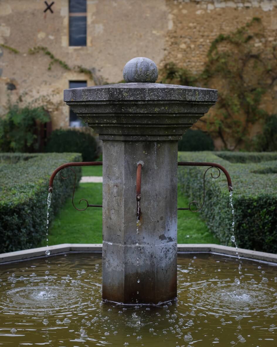 Stone fountain in the center of a garden pond surrounded by lush greenery and manicured hedges in Normandy, France.