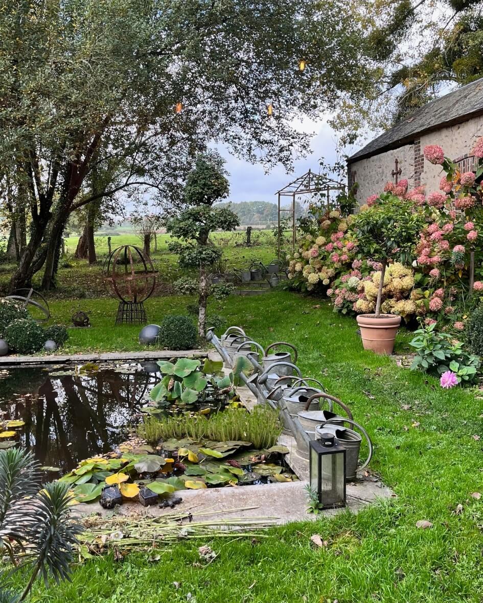 Lush outdoor garden in Normandy, France featuring pink hydrangeas and a pond lined by watering cans.