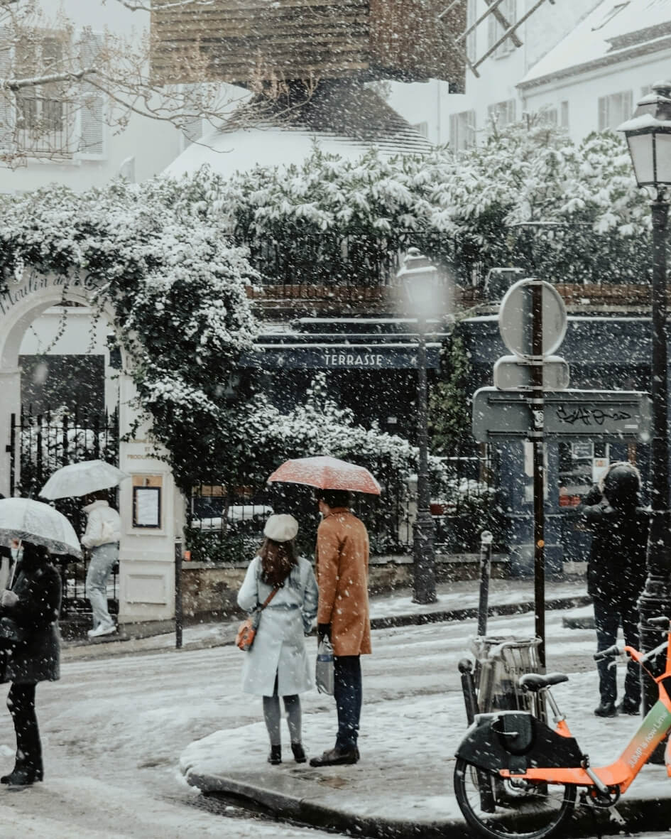 People strolling through snowy Parisian streets, bundled in warm coats and protected by umbrellas.