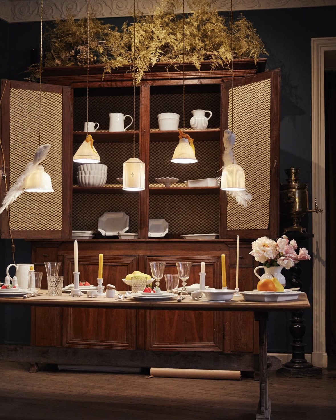 Dining room table set with cut flowers and candle sticks beneath decorative hanging light fixtures.