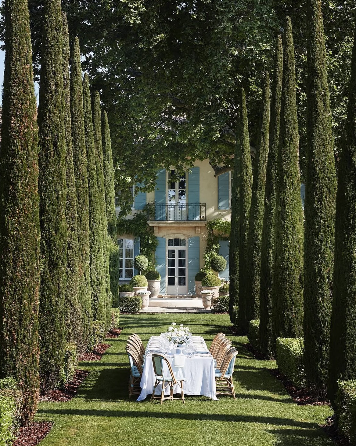 Dining table set among Cyprus trees in the garden of a luxury villa in Provence, France, with lush grass and bright natural light.