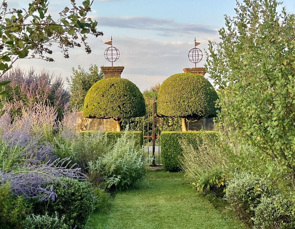 Lush garden path in Provence lined with lavender and flowering greenery, leading through iron gates and manicured trees.