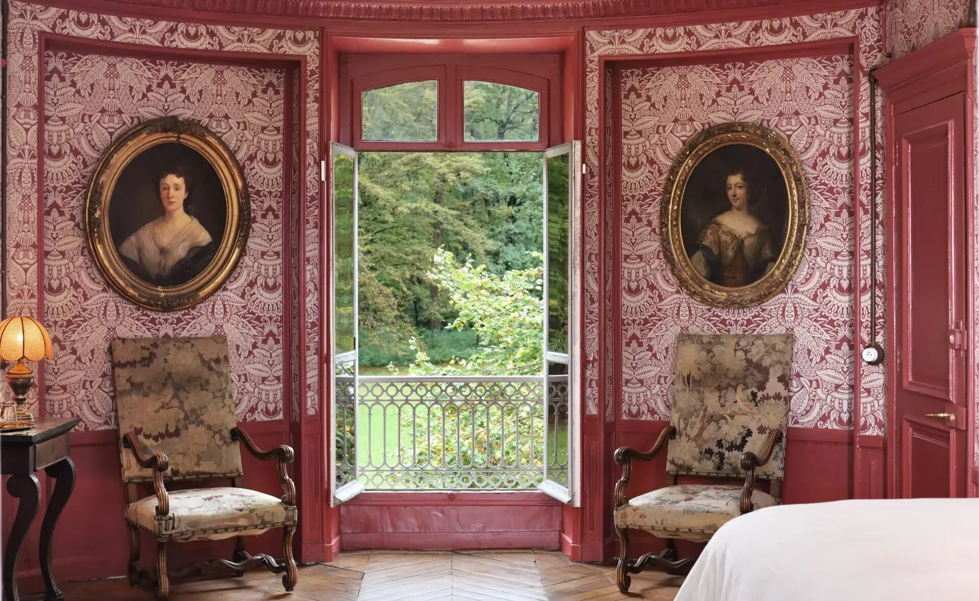 Chateau bedroom interior with a garden view featuring ornate red wallpaper, French antique furniture, and framed art.