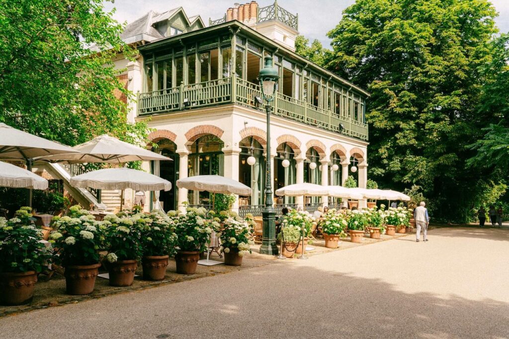 ORA Farm House restaurant exterior in Parc Buttes-Chaumont, Paris with a green painted sunroom, potted hydrangeas, and white umbrellas.
