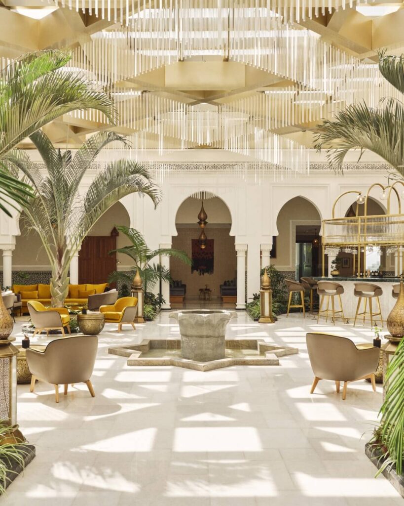 Four Seasons hotel interior in Morocco, featuring an open communal salon and bar with bright natural light, palm trees, and a stone fountain.