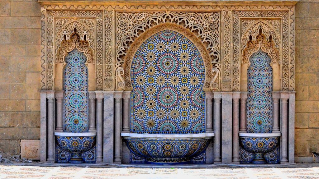 Ornate stone and mosaic Moroccan fountains with blue tiles displayed along a stone wall.