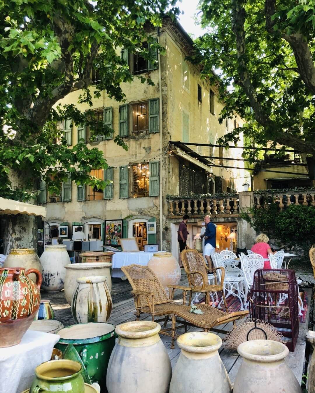 Open air antique market along the French Riviera featuring wicker chairs, large clay planter pots, and shoppers throughout.
