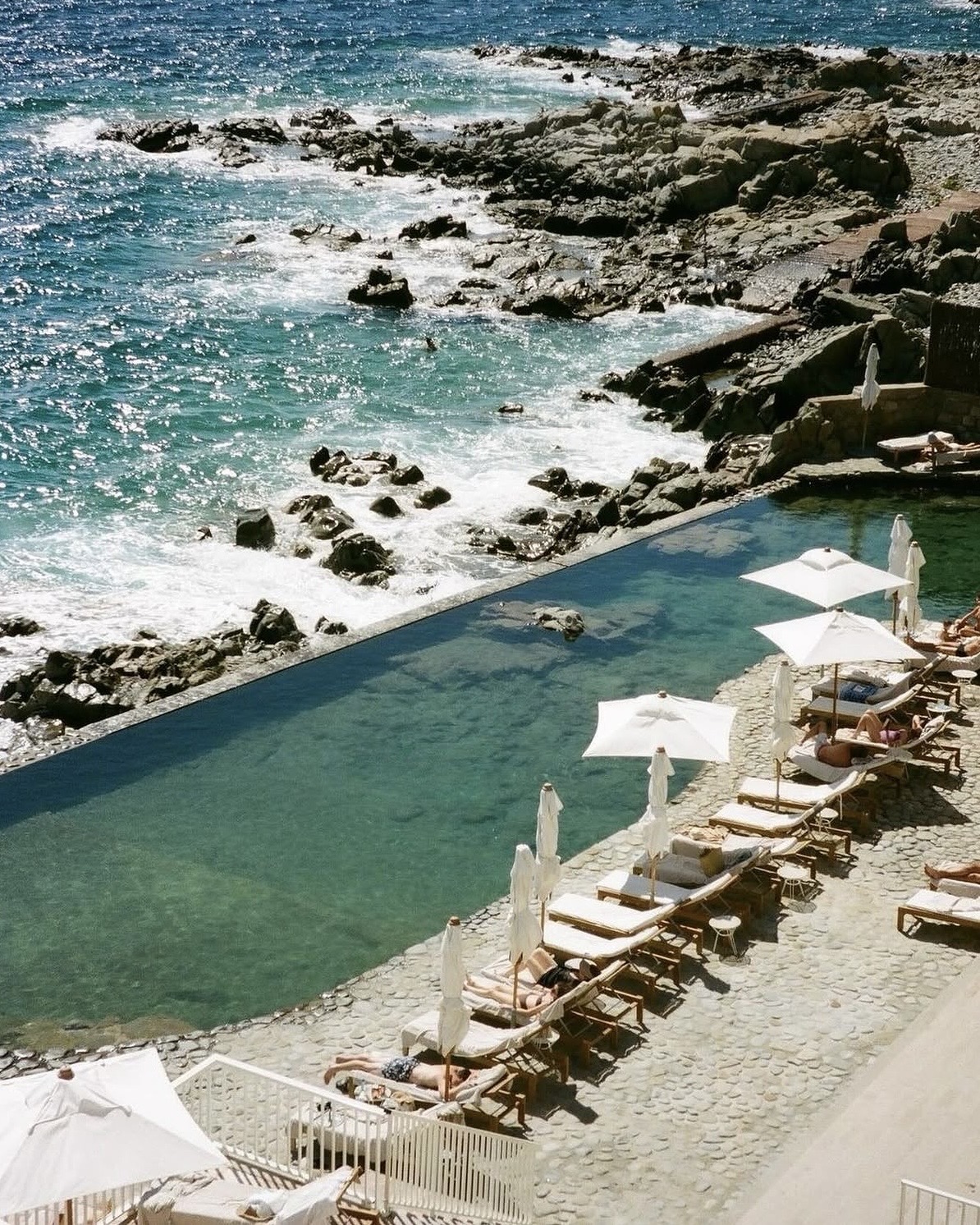 Les Roches Rouges luxury hotel exterior featuring a natural swimming pool lined with lounge chairs and umbrellas, beside the rocky coast.