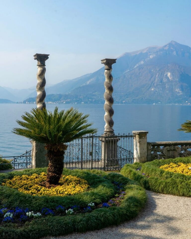 Ornate lush garden and iron gates along Lake Como with mountain views.