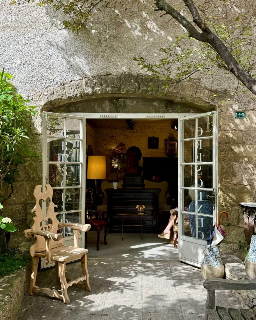 Antique store entrance with white French doors held open by antique furniture and a wooden chair in Provence.