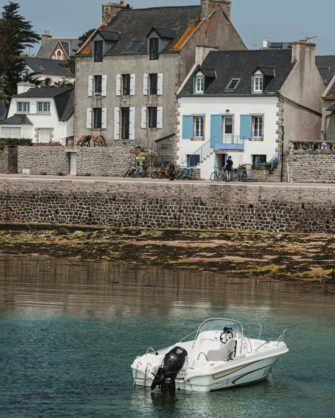 White boat moored along the Atlantic coastline of Île de Batz, framed by mossy rocks and French homes.