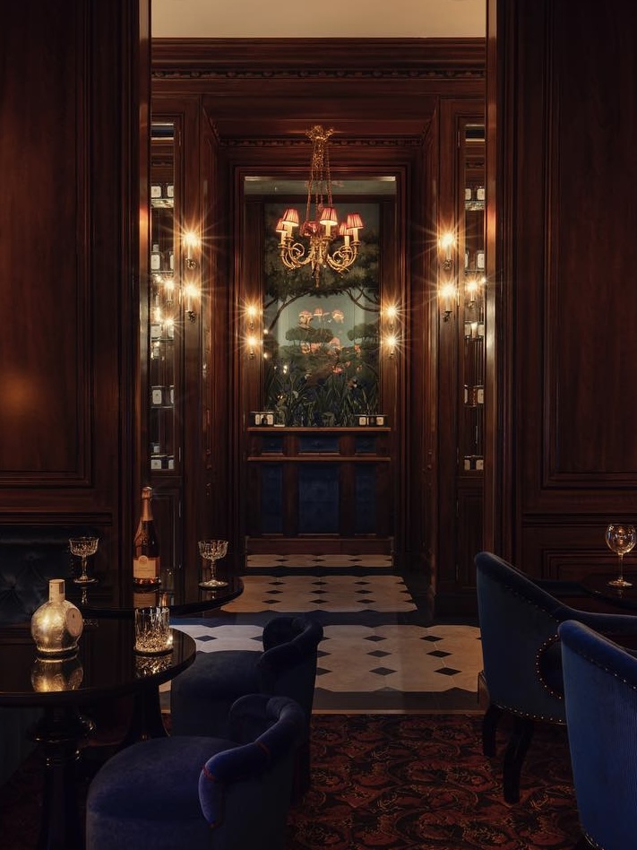 Maison Proust dimly lit hotel bar interior with velvet blue chairs, marble floors, and wood door frames.