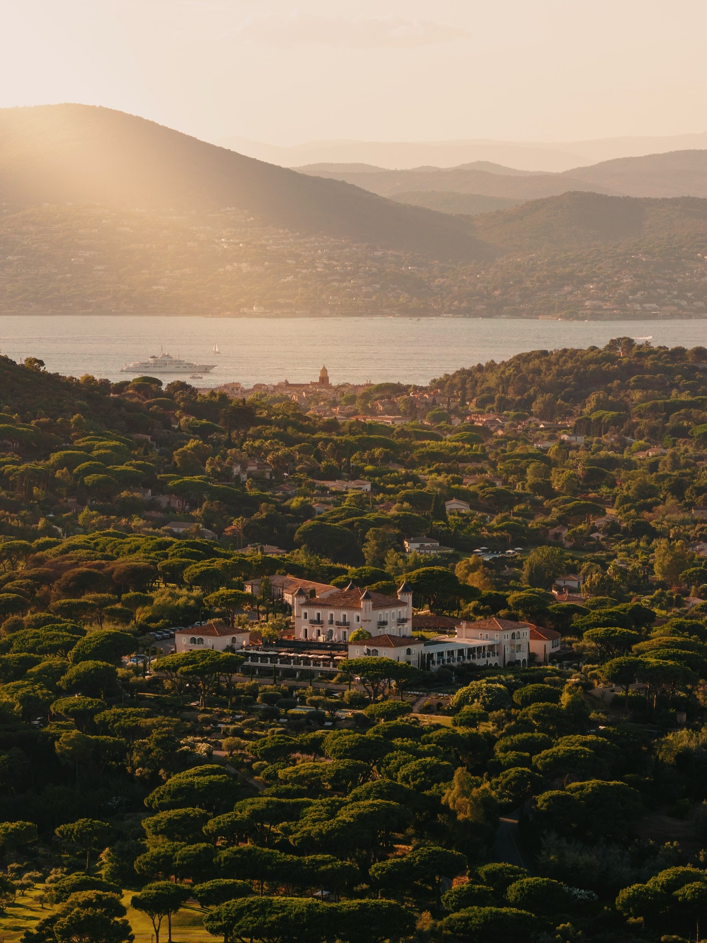 View of Château de la Messardière in Saint Tropez surrounded by lush green trees, the Mediterranean Sea and distant mountains.