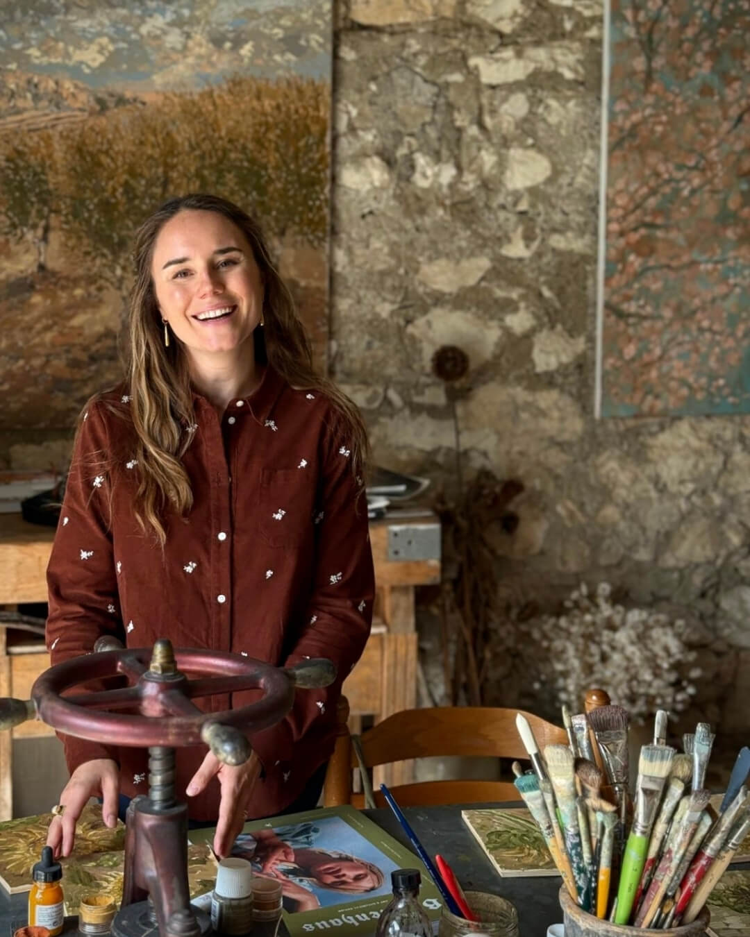 Ashley Tinker posing in an art studio surrounded by paintings, books, and paint brushes.