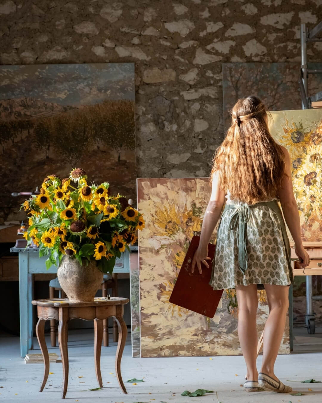 Ashley Tinker standing in her art studio surrounded by large paintings beside a vase of cut sunflowers.