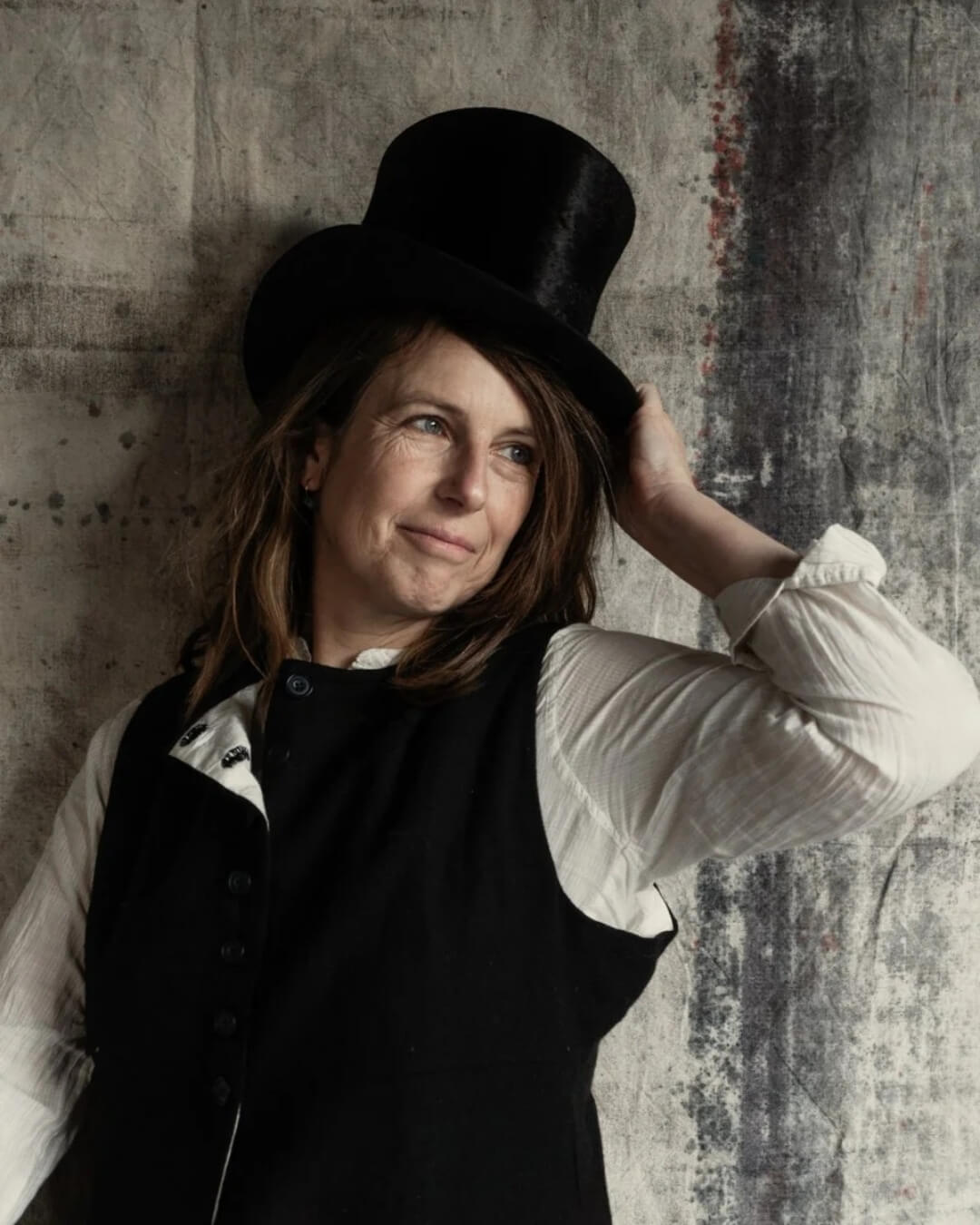 Joanna Maclennan posing against a rustic painted background wearing a suit and top hat.