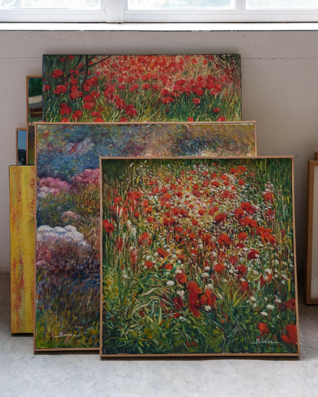 Floral and landscape paintings by Joseph Bayol arranged in a pile against a wall.