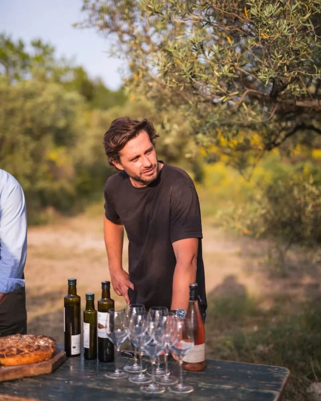 Man standing beside a garden table set with bred, oil, and wine in olive grove at Terres de Pierre in Provence.