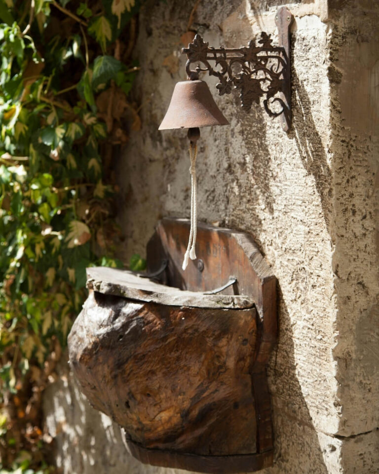 Rustic wood hutch fastened to a stone wall beneath a copper bell in a Provence garden.
