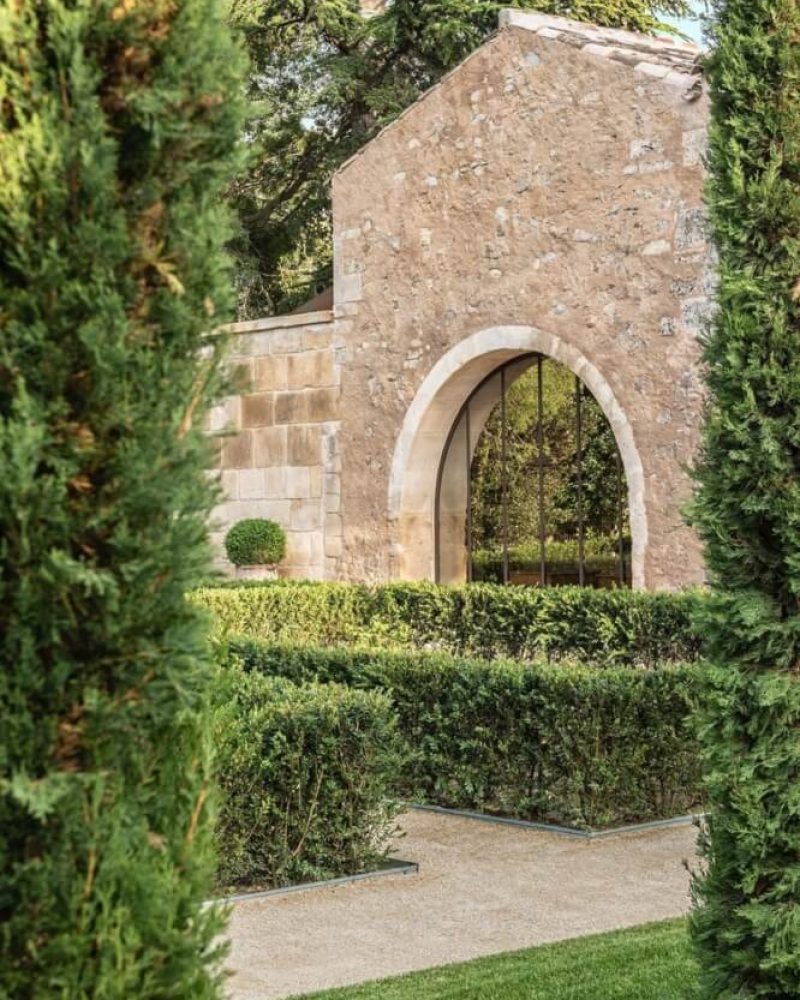 A pointed stone wall, lined with hedges and a pebbled garden walking path.